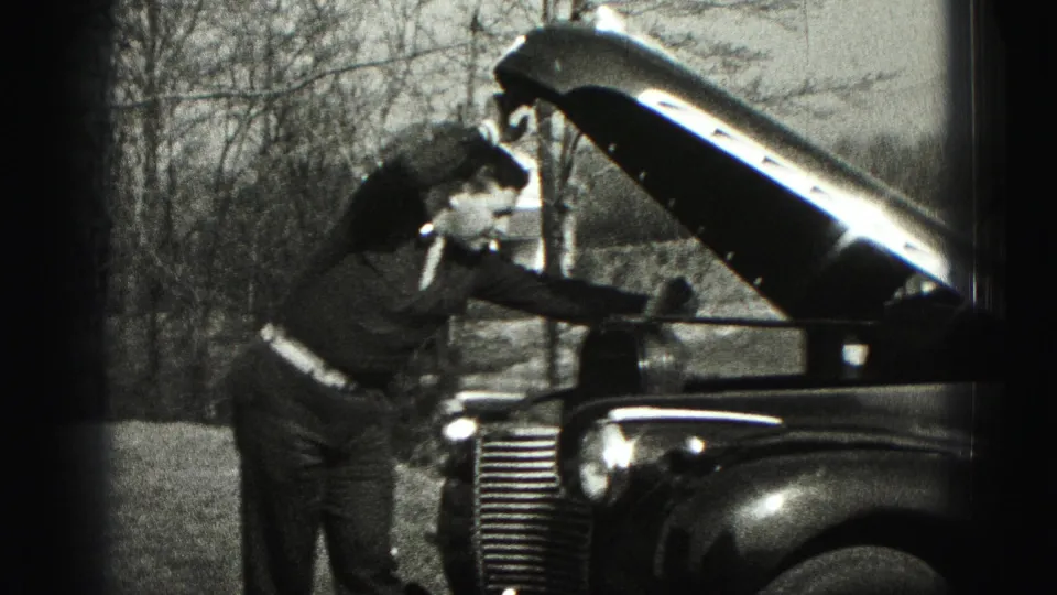 In Harrisburg, Pennsylvania footage from 1975, a man in a suit leans over the raised hood of a classic car outdoors.