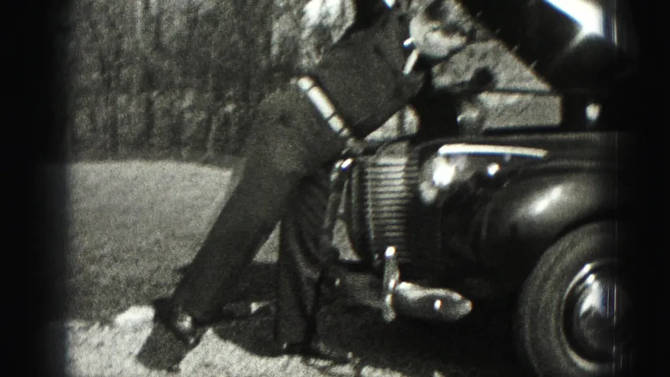 A suited man stands near the open hood and front fender of an older car in the 1975 Harrisburg home-movie footage.