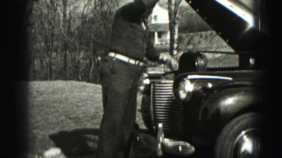 The man reaches toward the engine area under the raised hood of a vintage automobile in rural Harrisburg footage from 1975.