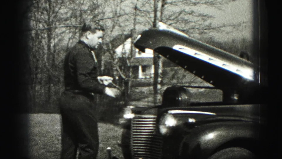 A man stands beside the open hood of a classic car, with trees and a distant house visible in 1975 Harrisburg home-movie footage.