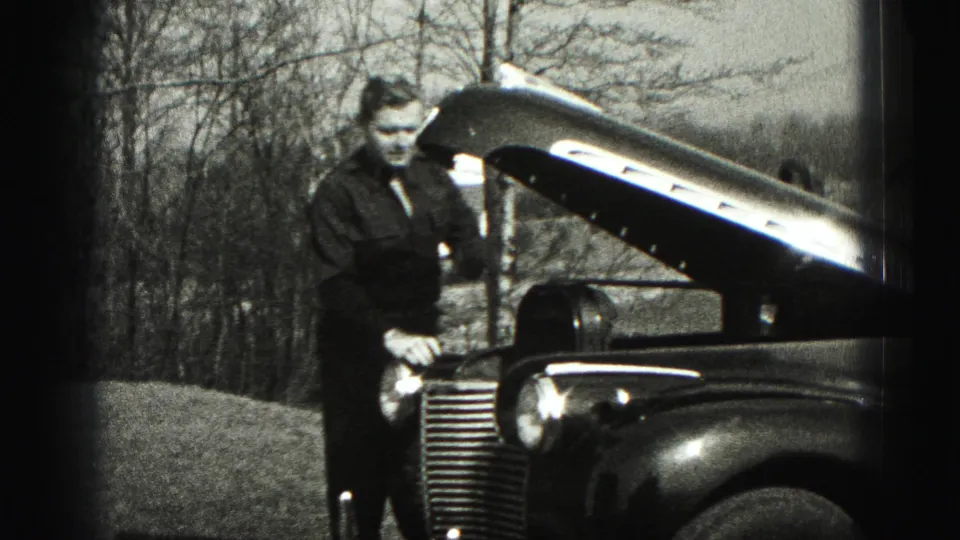 In 1975 Harrisburg home-movie footage, a man in dark clothing stands by a vintage car with the hood open outdoors.