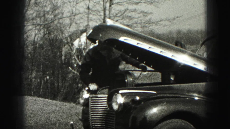 A man bends toward the engine area of a vintage car with its hood raised in 1975 Harrisburg home-movie footage.