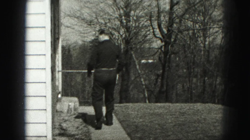 A man walks away from the camera along a narrow path beside a house exterior in Harrisburg home-movie footage.