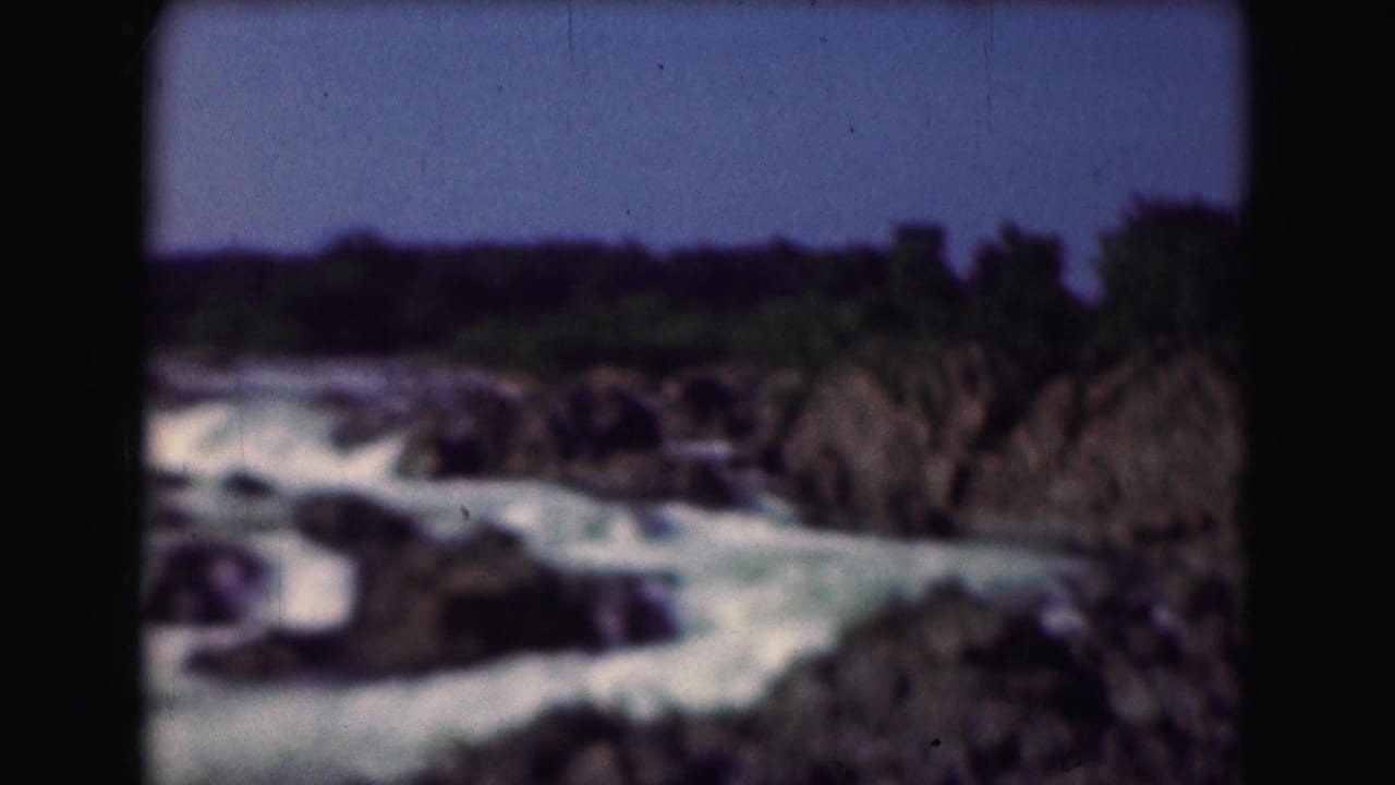 Rushing whitewater flows between dark rocks under a blue sky.