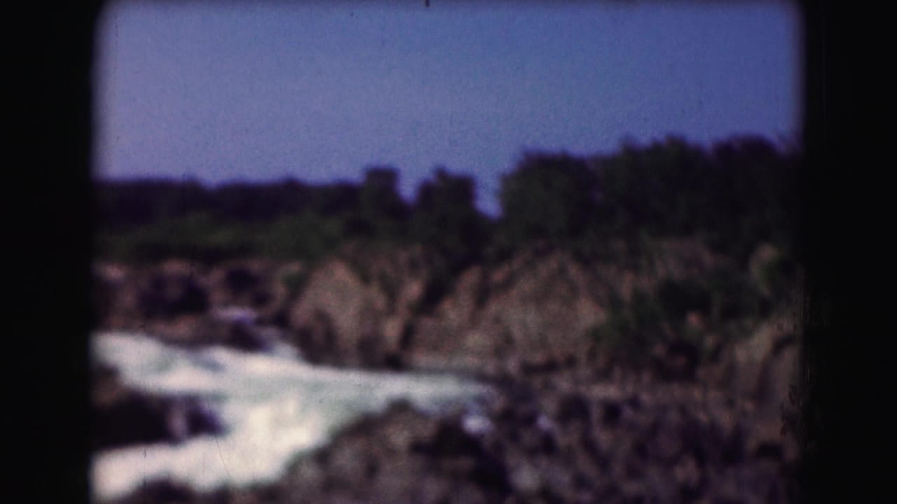 Soft 8mm view of a rocky waterfall and white seafoam, with low trees and blue sky beyond.