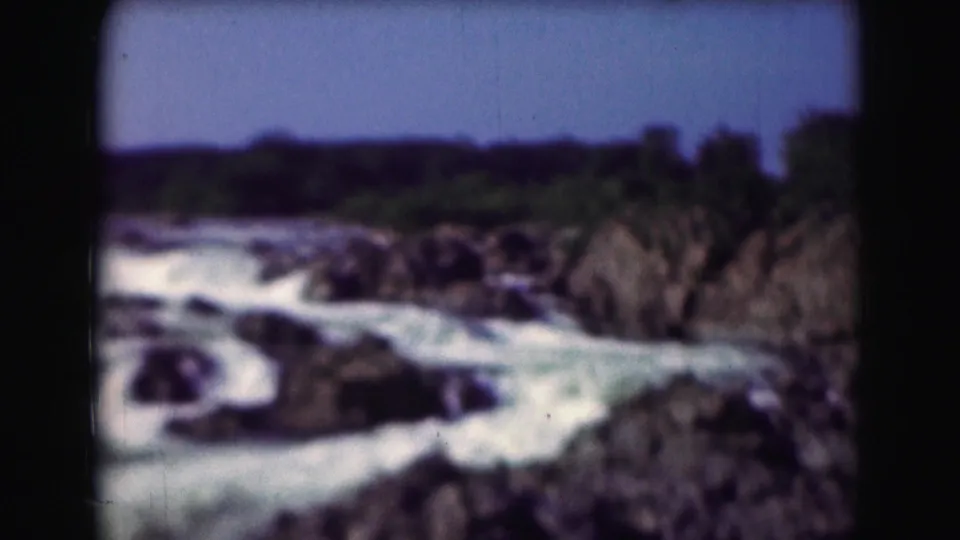 1975 home-movie frame showing foamy river water moving past dark rocks beneath a clear sky.