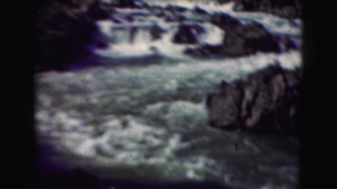 Aged 8mm view of turbulent blue-green water below small falls, bordered by dark rocky terrain.