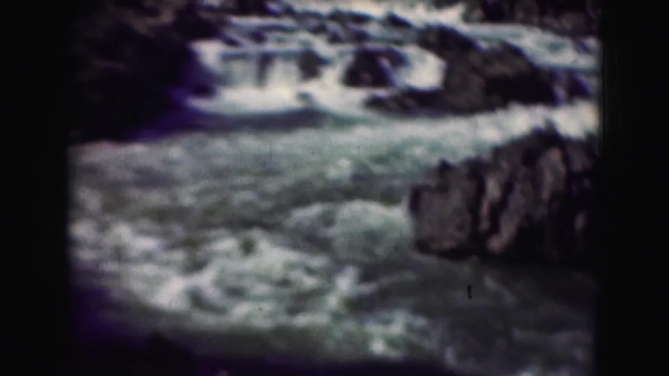 Aged 8mm view of turbulent blue-green water below small falls, bordered by dark rocky terrain.