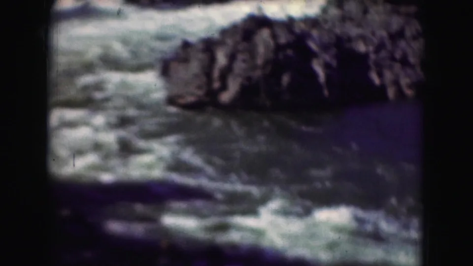 1975 home-movie frame showing foamy rapids moving past a large dark rock formation, with aged color shift.