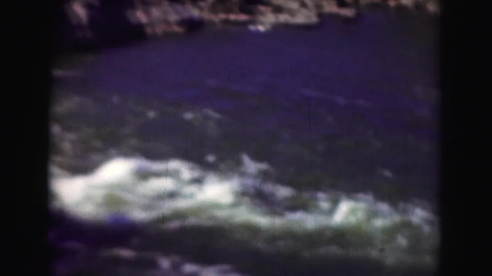 Soft 8mm frame of a blue river pool and foreground whitewater, with rocky terrain along the upper edge.