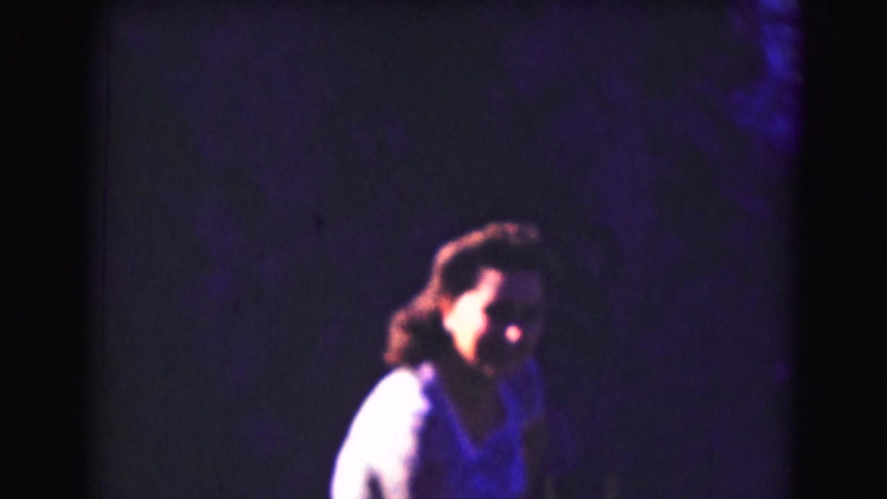 A person in a light-colored top appears in soft focus against a dark, purple-hued 8mm background during the 1975 home-movie sequence.