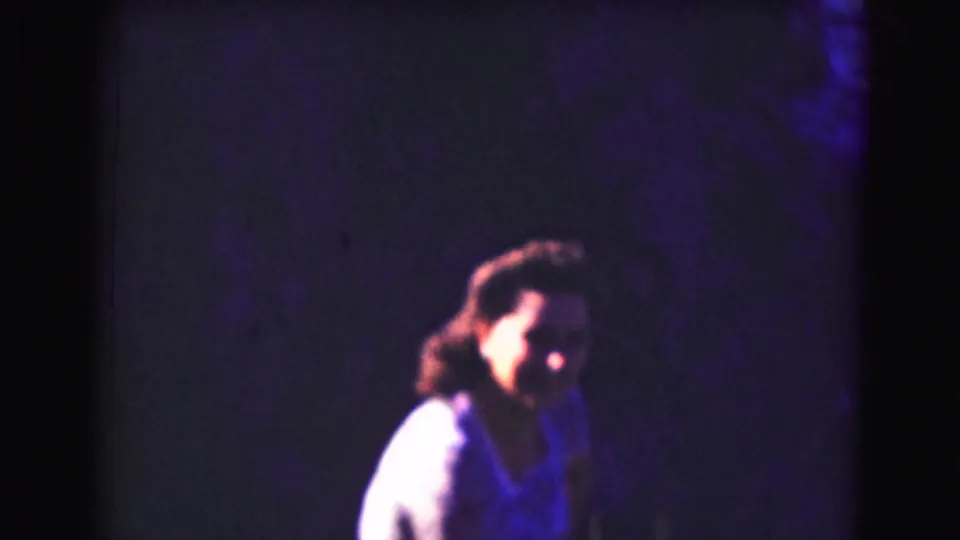 A person in a light-colored top appears in soft focus against a dark, purple-hued 8mm background during the 1975 home-movie sequence.