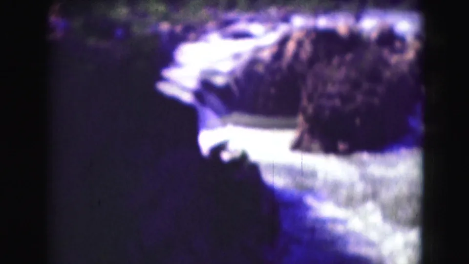 Soft-focus 1975 8mm view of whitewater rushing past dark rocks, with a shaded tree line beyond the river.