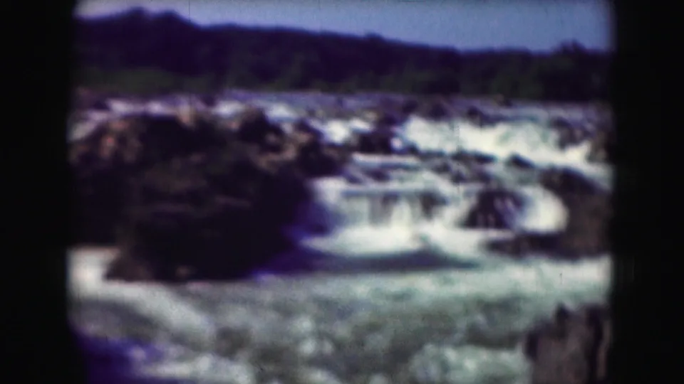 1975 Washington, DC-area 8mm frame showing rapids or small waterfalls flowing around rocks below distant trees and low hills.