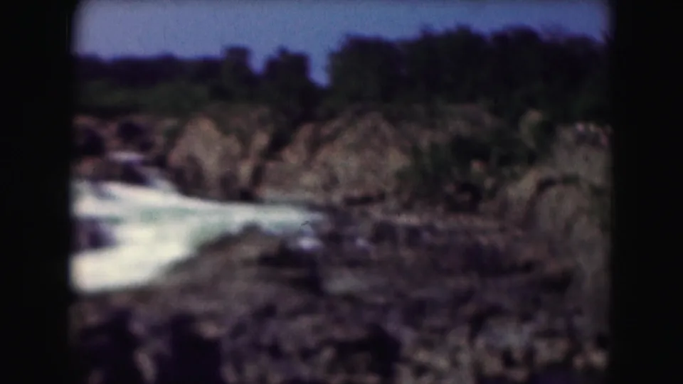 A 1975 8mm view of water, rocks, trees, and open sky in the Washington, DC area, framed with a handheld home-movie look.
