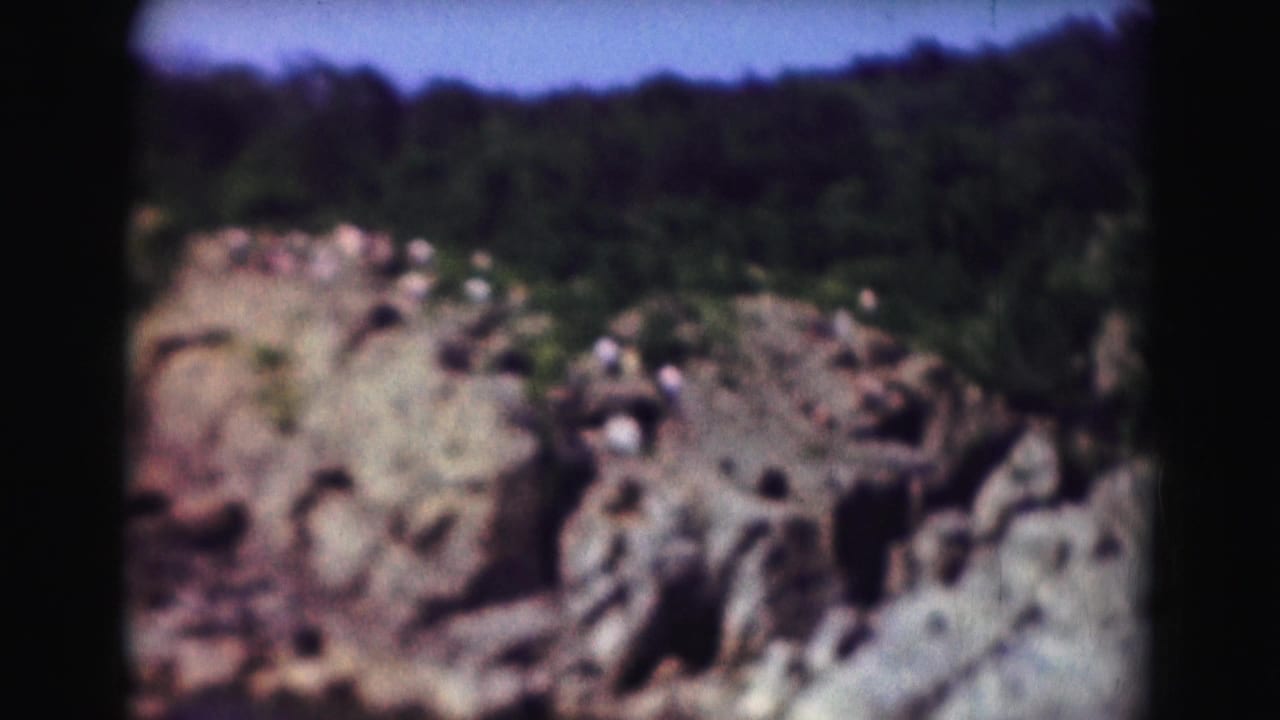 1975 8mm Washington DC footage showing a rocky river overlook with distant visitors, tree cover, and blue sky.