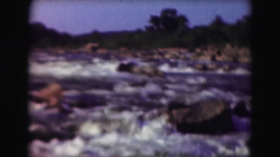 1975 8mm home-movie frame of river rapids churning around rocks, with a wooded bank in the background.