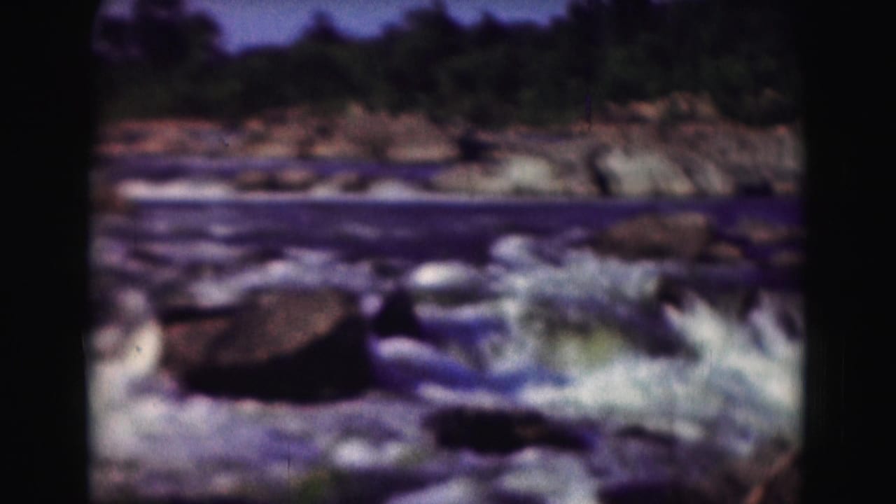 Soft-focus 1975 8mm view of fast-moving water and rocks, framed by dark film edges and distant trees.