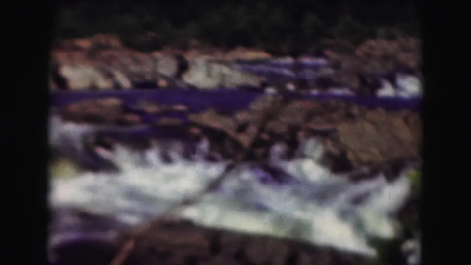 1975 8mm home-movie frame showing foaming river water breaking over dark rocks and ledges.