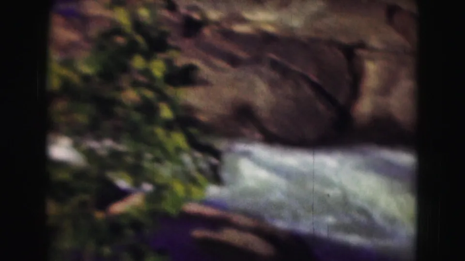 1975 8mm frame of river water passing near large smooth rocks and green vegetation along the bank.
