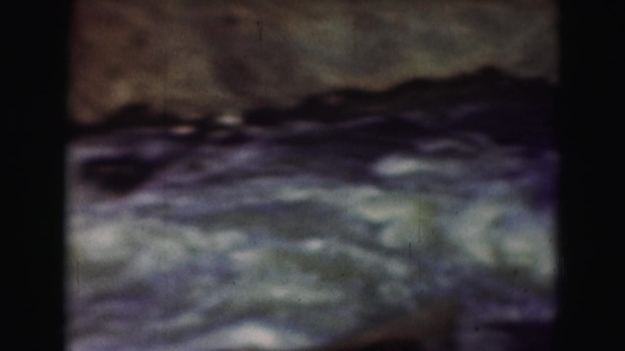 White water rapids continue past dark rocks in a soft, grainy frame.