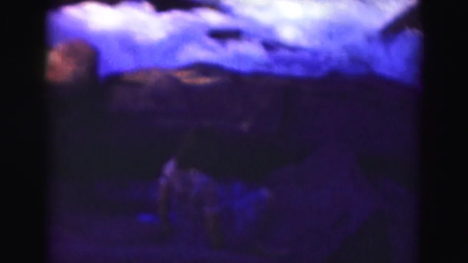 Blue-purple 1975 8mm home-movie frame near Washington, DC showing a teenage girl low on rocks beside moving water.