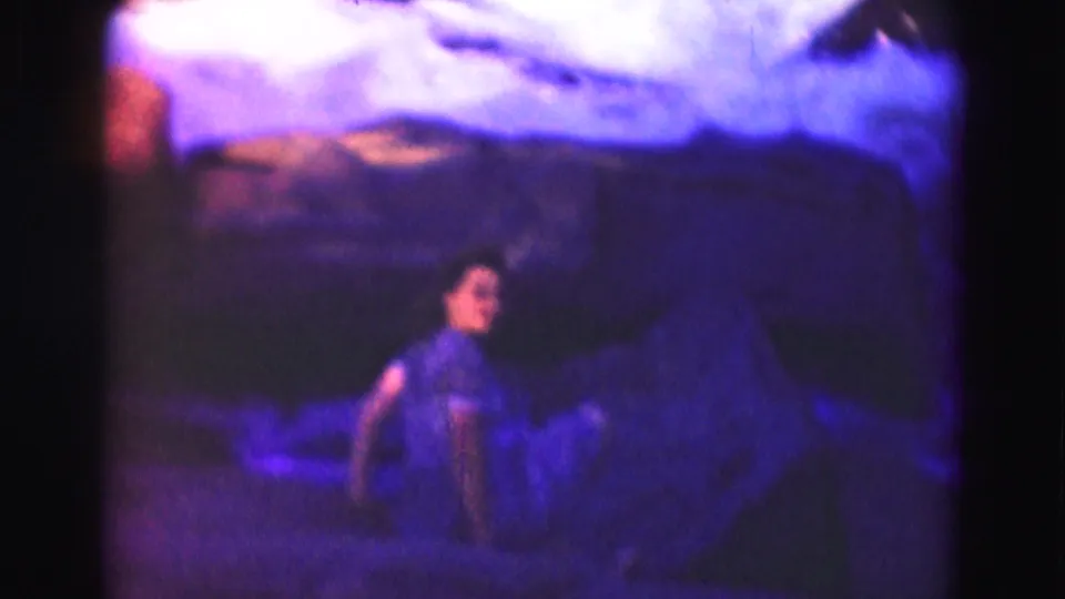 1975 8mm home-movie frame near Washington, DC showing a teenage girl crouched on rocks along a shaded river.