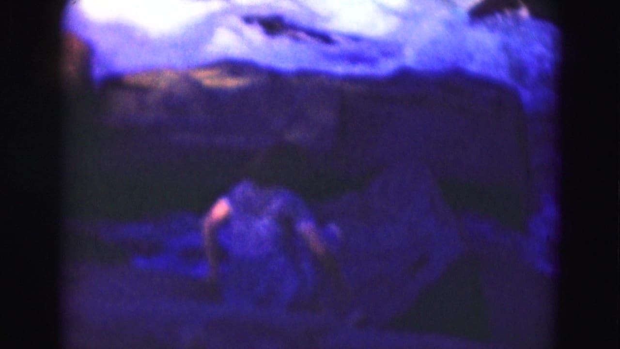 Soft, purple-toned frame of a figure near rocks and water under a cloudy sky.
