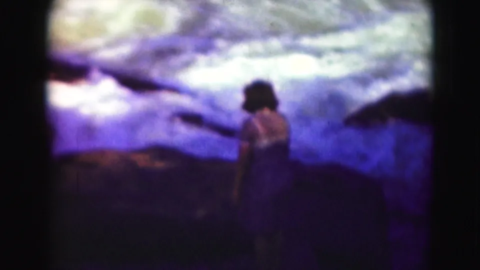 1975 8mm Washington, DC home-movie frame of a woman in a blue dress near rocks and water, seen from behind or in profile.