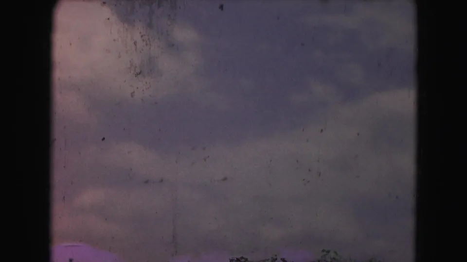 A faded 8mm frame shows layered clouds and faint aircraft shapes in the sky, with scratches and dust visible across the scan.