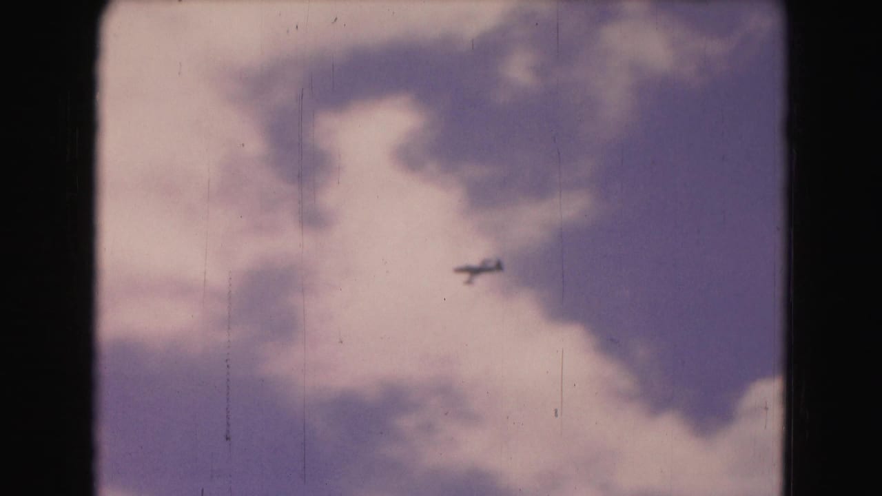 Small plane flying through a cloudy sky, framed by dark film edges.