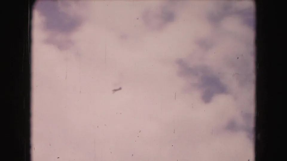 A small aircraft shape is visible against a pale, cloudy sky in the vintage 8mm footage.
