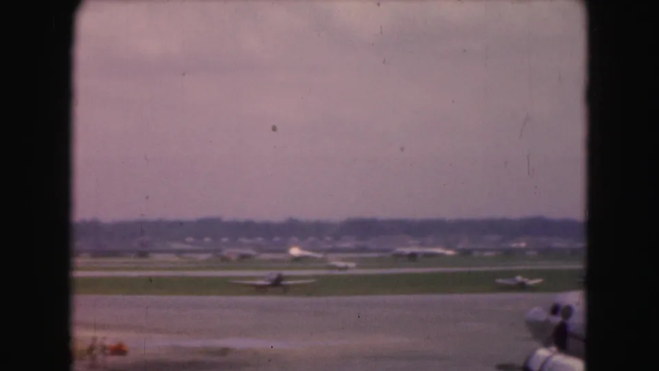 Washington DC airport runway view from 1975 8mm footage, showing planes on the ground, grass, pavement, and distant trees.