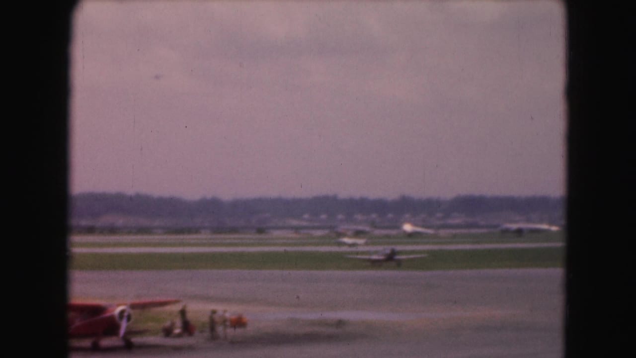 Small red biplane near a grass runway, seen through a dark window frame.