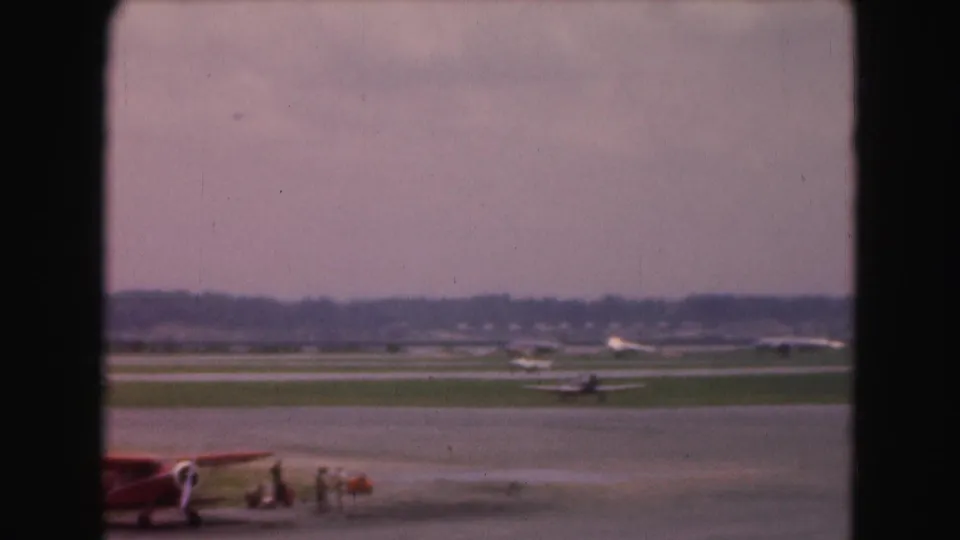 1975 Washington, DC-area airport view with a small red biplane and parked light aircraft seen through a window frame.
