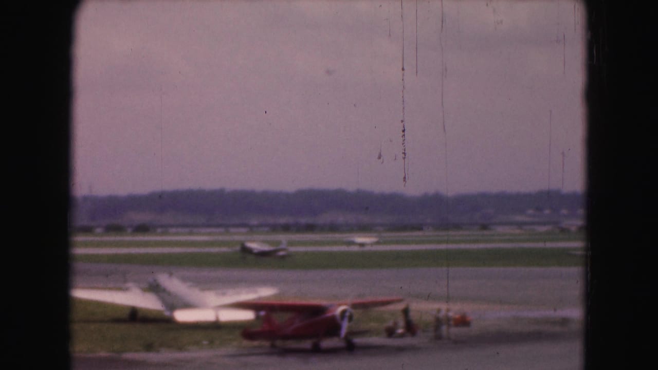 Small red biplane and nearby light aircraft on a grassy 1975 Washington, DC-area airfield, framed by dark vertical bars.