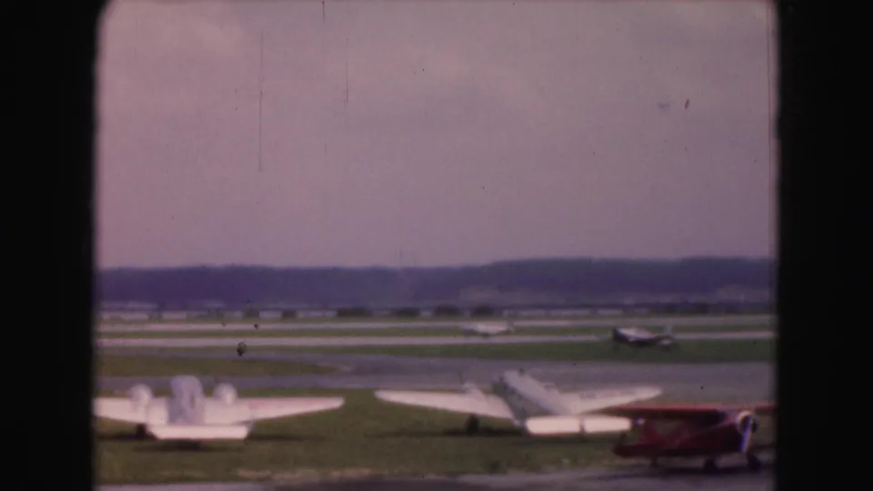 A white light aircraft and red biplane appear on a grass runway area in 1975 Washington, DC-area home-movie footage.