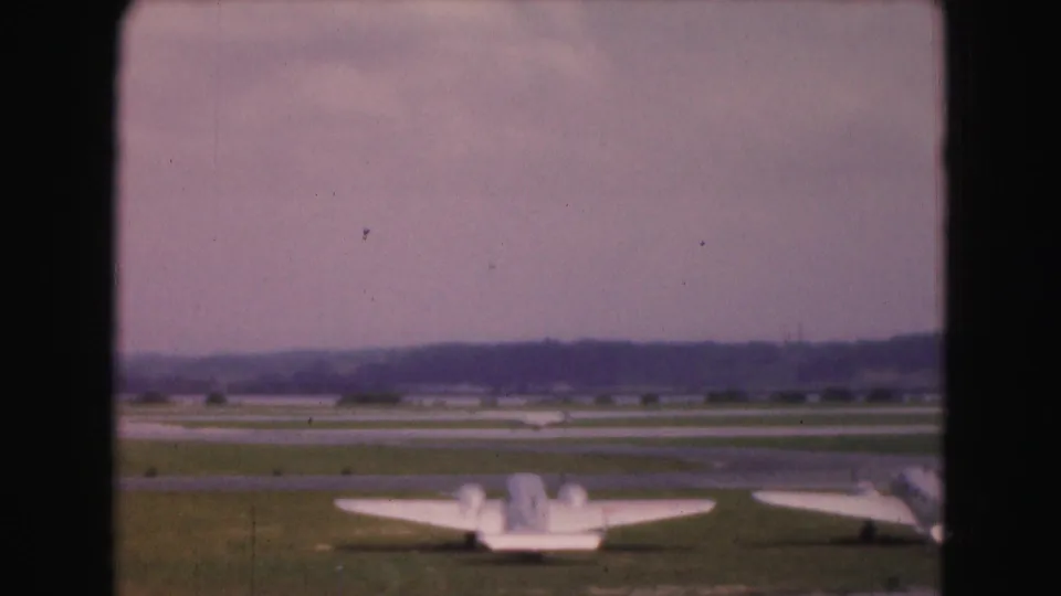 Wide 8mm view of small aircraft on a grassy Washington, DC-area airport field in 1975, with runway strips beyond.