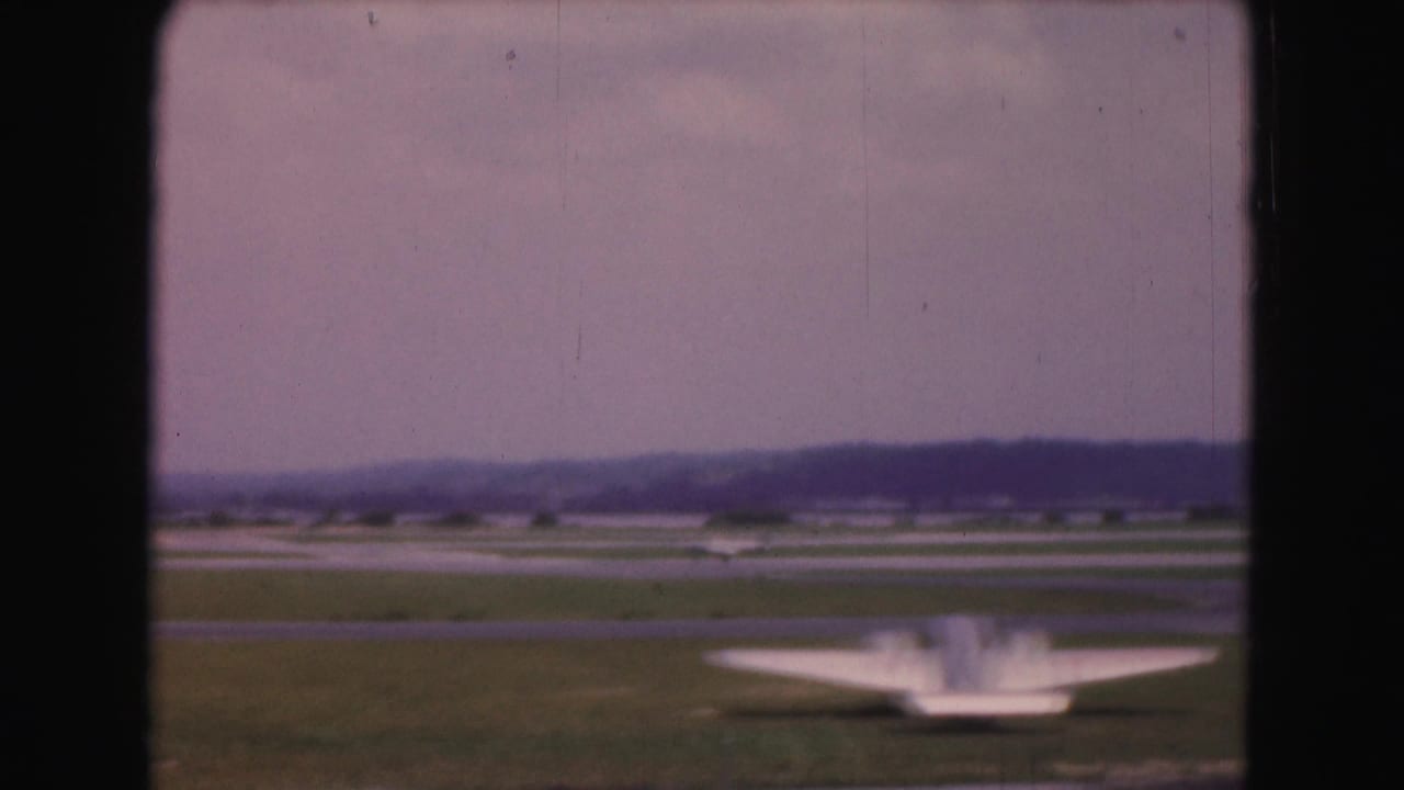 1975 Washington DC airport view through a window frame, showing a runway, grassy field, small aircraft, and hazy distant buildings.
