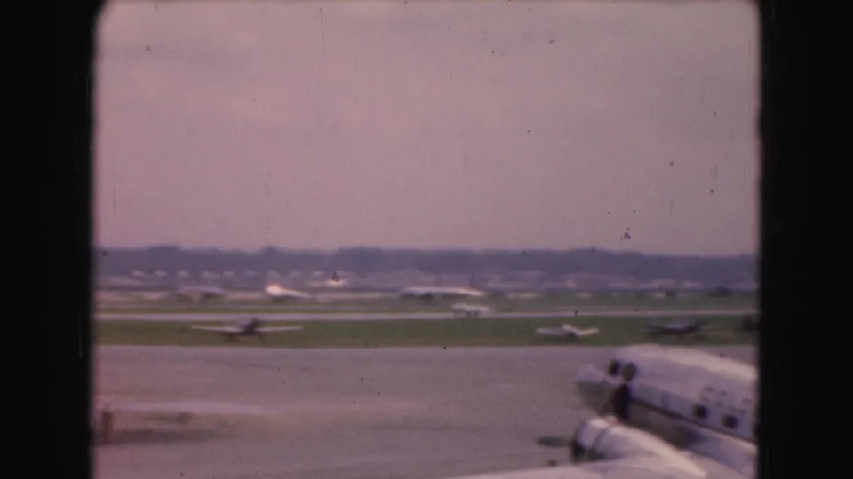 1975 Washington DC airport view showing a cropped airliner at the edge of frame, runway traffic, grassy strips, and distant aircraft.