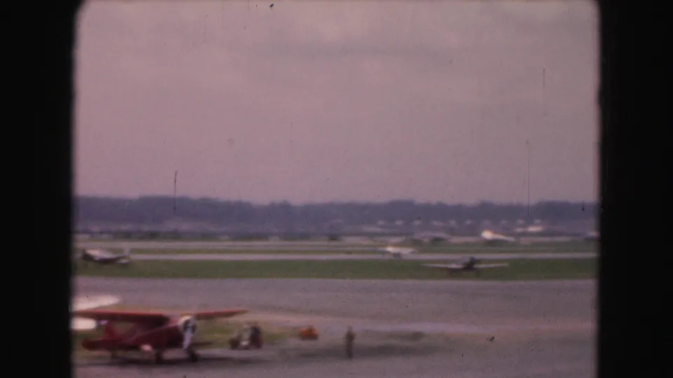 1975 Washington DC tarmac view with a small red aircraft, nearby figures, service items, runway, and distant airport traffic.