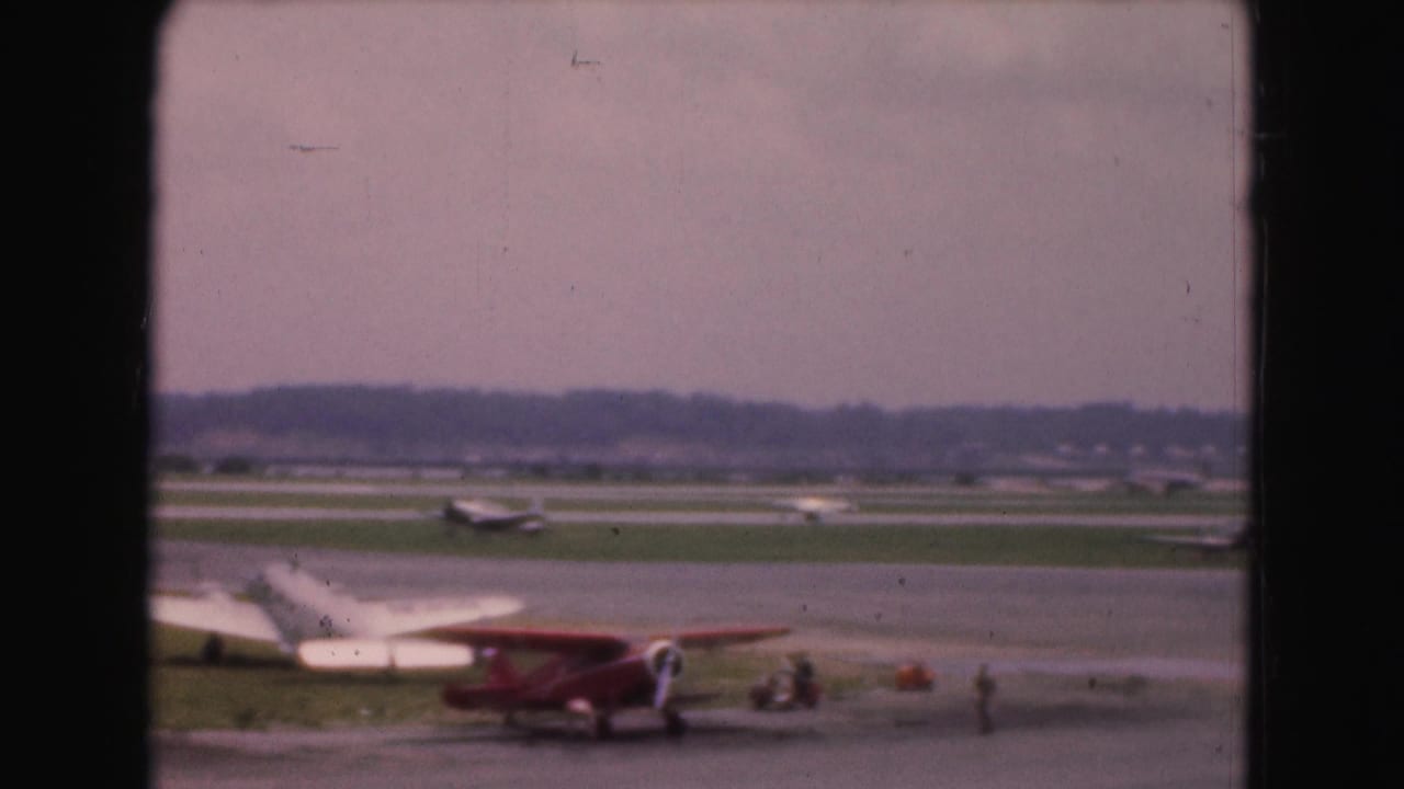 1975 Washington, DC-area home-movie view of a small airfield with light aircraft, runway grass, and a red biplane in the foreground.