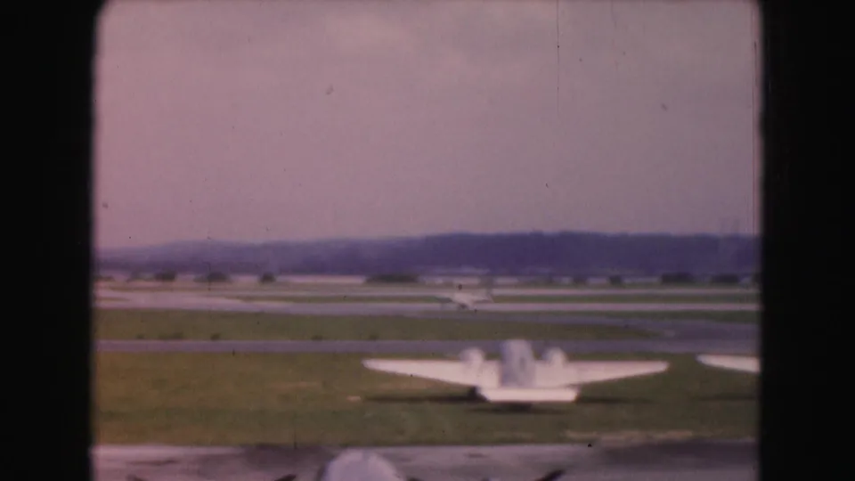 Soft 8mm view of a light aircraft from behind at a Washington, DC-area airfield, with runway grass and distant tree line visible.
