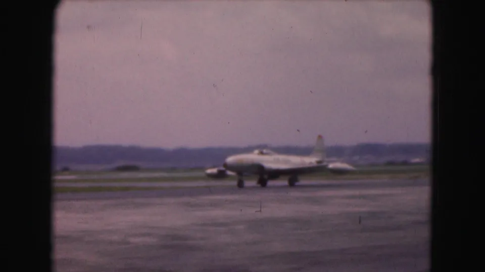 1975 Washington, DC-area airfield frame with a larger jet-like aircraft on the runway or taxiway under a cloudy sky.