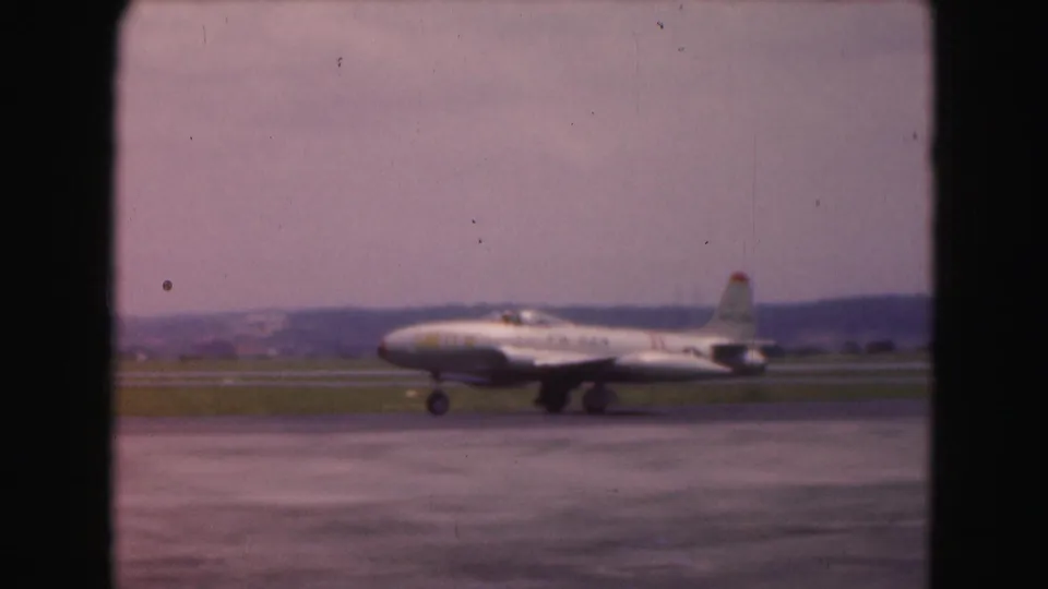 A white jet passes left to right on the runway, with soft focus and purplish 8mm color from the 1975 airport scene.