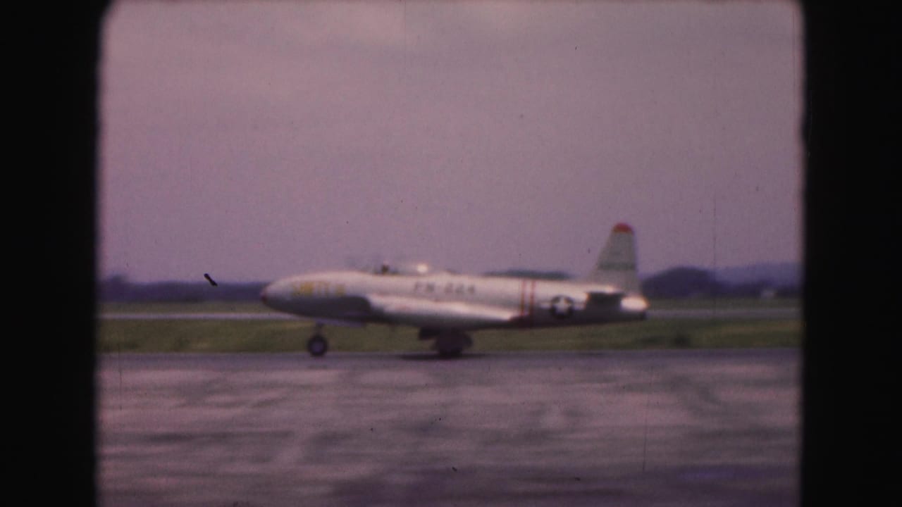White military-marked jet taxiing on a runway beside grass and distant trees.