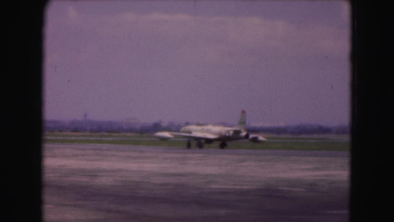 Silver military jet taxiing on a runway with grass and distant airfield background.