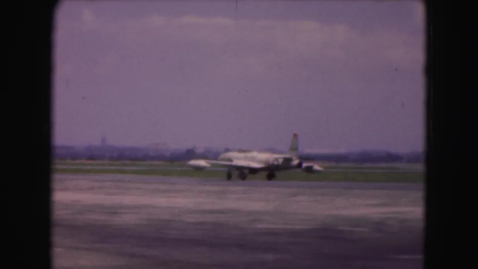 1975 8mm frame from Washington, DC metadata showing a silver military jet taxiing across a runway at a distance.
