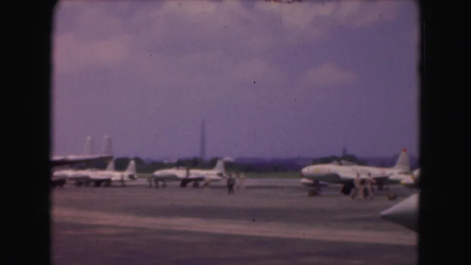 Wide 1975 8mm Washington DC airfield view with several parked military jets and people walking near the aircraft.