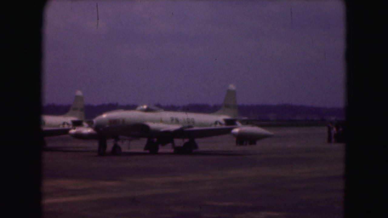 A silver military jet appears parked on the tarmac at the 1975 air base, with additional aircraft in the background.